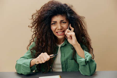 Irritated stressed angry tanned curly Latin lady talking with phone sit at the table squeezes pen...
