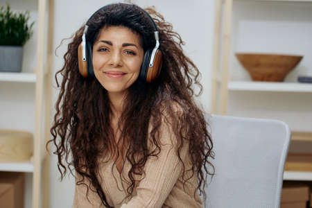 Smiling cute tanned curly Latin lady with headphones enjoy cool playlist listen music sit on chair in home modern interior look at camera. Copy space Mockup Banner. Concept sound stream. Home relaxの写真素材