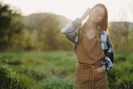 Portrait of a young smiling woman in work clothes checkered shirt and apron in nature in the evening after workの写真素材