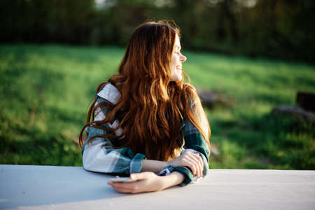 Girl with phone in hand sitting in nature in the park at the table smiling beautifully and looking...