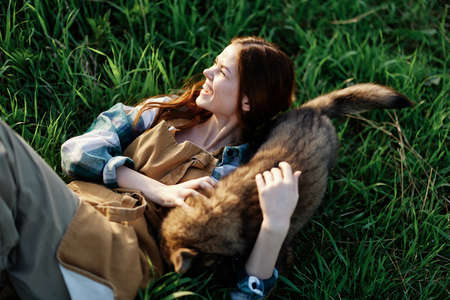 A woman lies on the grass smiling and cuddling her dog in nature in a park in the summer sunset. The...