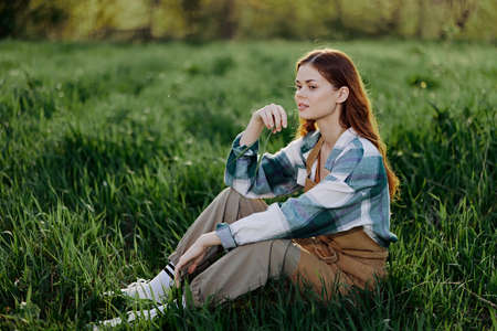 A young beautiful woman sits on the green grass in the park and looks out into the setting sunの写真素材