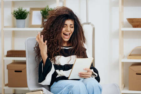 Overjoyed angry concentrated pretty beautiful curly tanned Latin woman in striped shirt hold notebook look down read work letter essay in home interior. Copy space Banner. Writer conceptの写真素材
