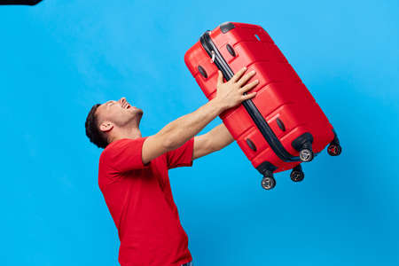 Overjoyed excited happy tanned handsome man in red t-shirt preparing for holiday hold big suitcase posing isolated on blue studio background. Copy space Banner Mockup. Trip journeys conceptの写真素材