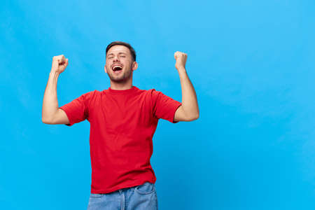 Overjoyed happy tanned handsome man in red t-shirt raise fist up have big win say Yeah posing isolated on blue studio background. Copy space Banner Mockup. People emotions Lifestyle conceptの写真素材