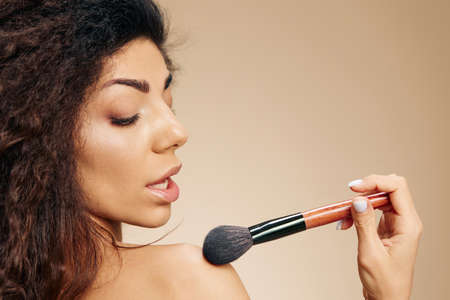 VISAGE CONCEPT. Profile sexy adorable tanned awesome curly Latin lady hold makeup brush on shoulder apply powder highlighter posing isolated on pastel beige background look down. Copy space bannerの写真素材