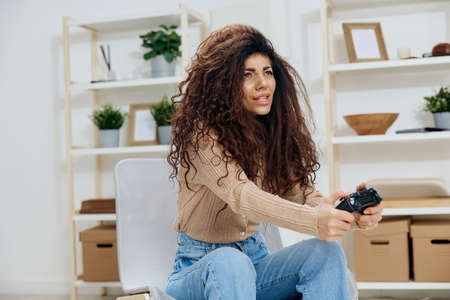 Overjoyed happy tanned curly Latin winner lady hold with joystick gamepad in both hands smile look aside sit on chair in home modern interior. Copy space Mockup Banner. Concept game platformの写真素材