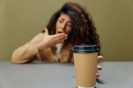 Tired sleepy tanned curly Latin female yawn hold for a cup to-go to enjoy coffee sit at the table isolated over olive green background. Copy space Mockup Banner. Coffee lover offer. Wide angleの写真素材