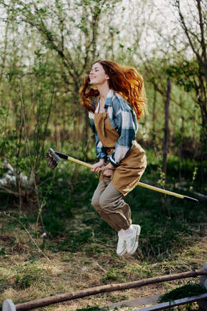 The woman hops amusingly with her rake like a witch on a broomstick in her green garden among the...