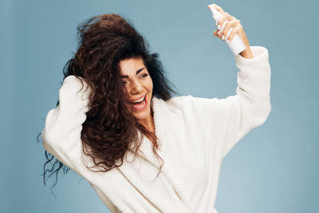 Hairstyling morning routine. Overjoyed tanned curly Latin lady in bathrobe Spraying On Hair For Repair split ends posing isolated on pastel blue background, smiling aside. Copy space offerの写真素材