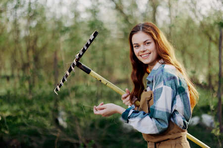 A woman smiling beautifully and looking at the camera, a farmer in work clothes and an apron working outdoors in nature and holding a rake to gather grass and forage for the animals in the gardenの写真素材