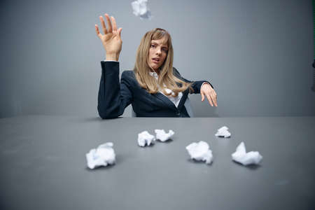Funny millennial blonde businesswoman worker in blue jacket throws papers into the camera sitting at...