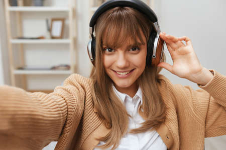 Cute young blonde student lady freelancer in warm sweater in headphones listen fav songs sitting in...