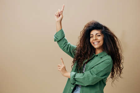 LOOK AT THIS CONCEPT. Smiling tanned curly Latin female in casual things point finger up posing isolated over pastel beige background look at camera show copy space Mockup Banner. Good offerの写真素材
