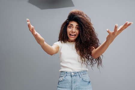 Close up shot of surprised terrific young lady stares with eyes popped out, notices something terrible in front, spreading hands at camera, posing isolated at gray background. People emotions conceptの写真素材