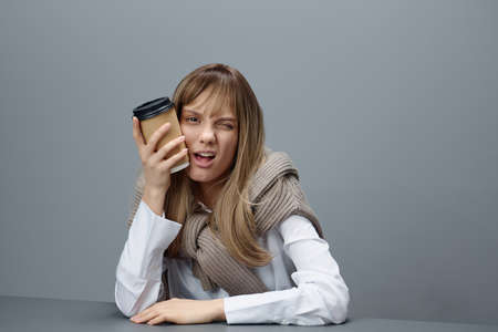 Winking happy cute young blonde student lady freelancer in warm sweater with takeaway coffee sitting...