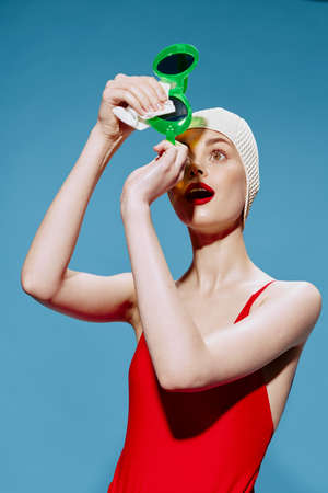 An enthusiastic shocked unusual girl in a swimming cap and a red swimsuit wipes glasses in the studio on a blue backgroundの写真素材
