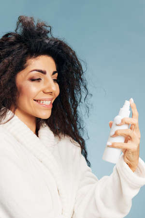 Smiling curly Latin lady in bathrobe look at toner spray in hand posing isolated on pastel blue background, feeling excited of effective skincare routine. Natural cosmetic ad concept. Copy spaceの写真素材