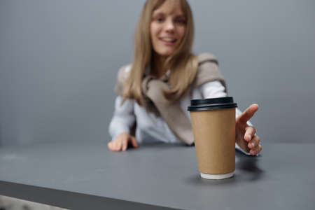 Happy young blonde student lady in warm sweater pulls hand with takeaway coffee at camera sitting at...