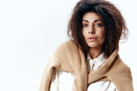 Smiling Calm Curly Pretty Latin Woman in White Shirt Touching Soft Pastel Beige Warm Sweater Posing Isolated Look At You At White Studio Wall Background. Fashion Huge Sale New Collection Conceptの写真素材