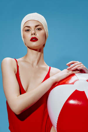 Retro fashion concept. A self-confident pretty lady in a swimming cap a red swimsuit with a ball on a blue background looks at the cameraの写真素材