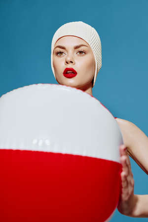 Focused woman in a red swimsuit swim cap with a striped ball looks ahead posing on a blue...