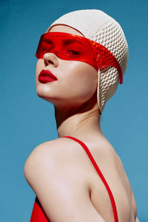 A self-confident stylish young woman in a white swimming cap and red glasses looks at the camera arrogantly. Headshot in the studioの写真素材