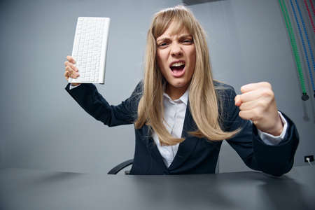 Crazy angry millennial blonde businesswoman worker in blue jacket throws keyboard and threatens at camera with fist in gray modern office. People Emotions Business Concept. Copy space, wide angleの写真素材
