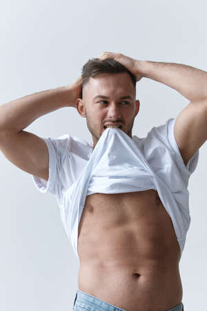 Portrait shot of handsome sexy flirting tanned man guy biting basic t-shirt touch hair posing on white background. Fashion Style New Collection Offer. Copy space for ad. Model snapの写真素材