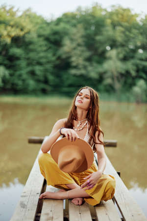 A young woman smiling in an image of a hippie and eco-clothes sitting outdoors on a bridge by a lake wearing a hat and yellow pants in the summer sunsetの写真素材