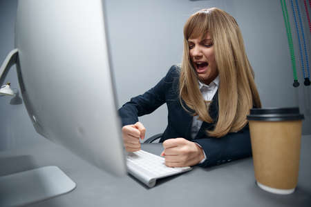 Evil irritated millennial blonde businesswoman worker in blue jacket breaks keyboard hate operation crash in gray modern office. Remote Job, Technology And Career Profession Concept. Wide angleの写真素材