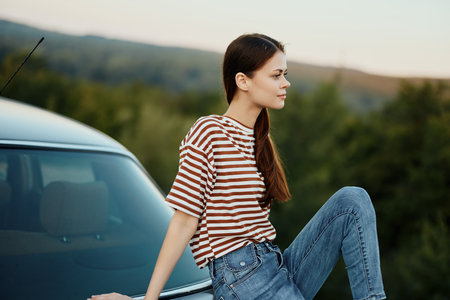A young woman traveler sits on the trunk of her car and watches the sunset in nature. The concept of freedom and travel as a lifestyleの写真素材