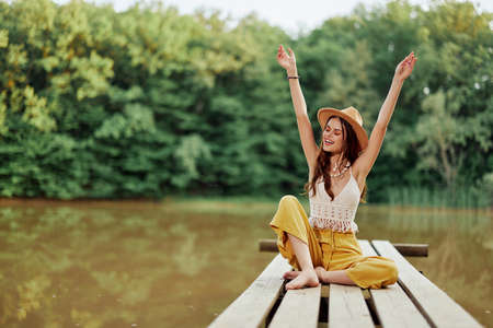 Hippie eco-activist woman traveler sits on a bridge by a lake with her arms outstretched with a hat and smiling sincerelyの写真素材