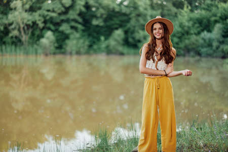A young woman in a hippie look travels in nature by the lake wearing a hat and yellow pants in the...