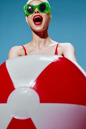A screaming cheerful unusual lady in a swimming cap pulls a red white ball into the camera on a blue background. Time to go to the seaの写真素材