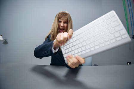Crazy irritated angry millennial blonde businesswoman worker in blue jacket throws keyboard and screaming at camera in gray modern office. People Emotions Business Concept. Copy space, wide angleの写真素材