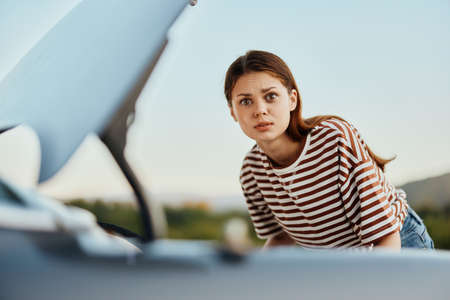 A sad woman looks in disbelief at her broken down car on the road while traveling and does not know...