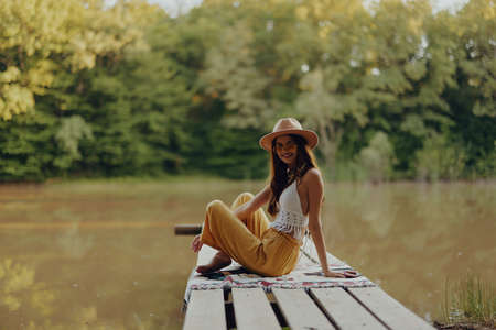 A hippie woman sits on a bridge by the river and enjoys the beautiful scenery around herの写真素材