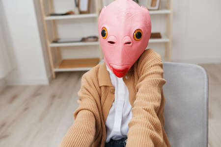 Cute funny blonde lady in pink fish mask warm sweater doing selfie use phone sitting in armchair at modern home interior. Pause from work, take a break, social media in free time concept. Wide angleの写真素材