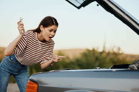 A woman with a wrench with a smile looks happily under the open hood of her car and repairs it from a roadside breakdown while traveling alone in natureの写真素材