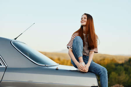A woman car driver sits on the trunk of a car and smiles admiring a beautiful view of autumn nature and mountainsの写真素材