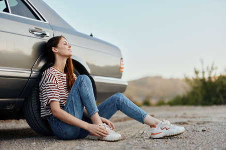 A young woman sits on the ground near her car on the side of the road and looks at the sunset. Camping after a hard road tripの写真素材