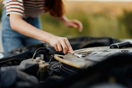 A woman traveler stands by her old car with the hood open and repairs the car engine with a wrenchの写真素材