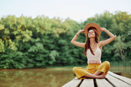 Hippie eco-activist woman traveler sits on a bridge by a lake with her arms outstretched with a hat and smiling sincerelyの写真素材