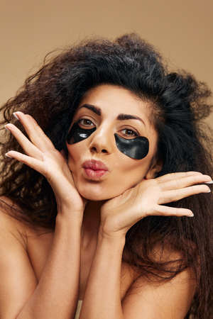Cute happy pretty Latin female with black hydrogel patches under eye blow kiss posing isolated over...