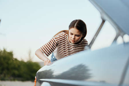 A woman traveler stands by her old car with the hood open and repairs the car engine with a wrench, unscrewing car partsの写真素材