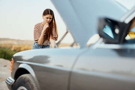 A woman traveler stands by her old car with the hood open, looking for the cause of the car...