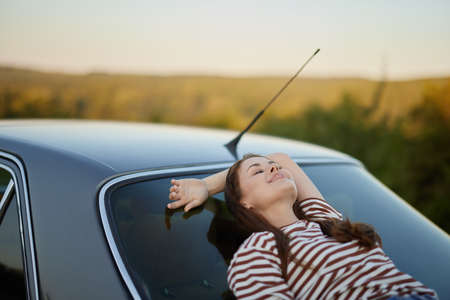 The woman driver stopped on the road and lay down on the car to rest and look at the beautiful landscape in a striped T-shirt and jeans. Complicated journey to natureの写真素材
