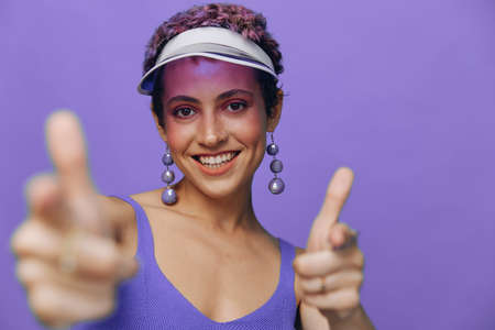 Portrait of a sporty fashion woman posing smiling with teeth and pointing a finger at the camera in a purple yoga tracksuit and a transparent cap on a purple monochrome backgroundの写真素材