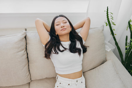 A young Asian woman sits at home on the couch resting asleep with her hands behind her head and smiling, waking up in the morning and drowsy during the dayの写真素材